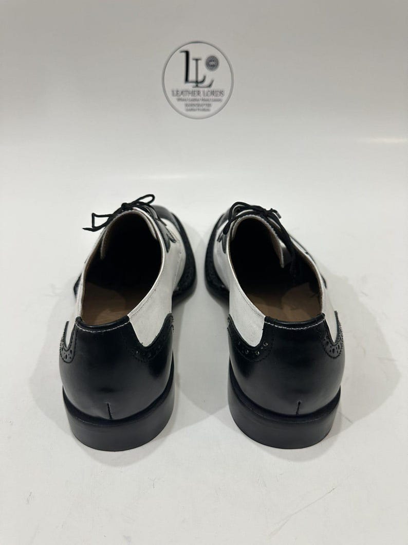 May include: A pair of black and white wingtip oxford shoes featuring a two-tone leather design, black laces, and decorative brogue detailing on the heel counters. The shoes are positioned with the heels facing forward, displaying a stacked heel.