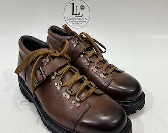 May include: A pair of brown leather boots with a rounded toe, thick black lug soles, and brown laces. The boots feature a side buckle strap, metal eyelets, and a logo on the upper ankle area that reads LEATHER LORDS Where Leather Meets Luxury.