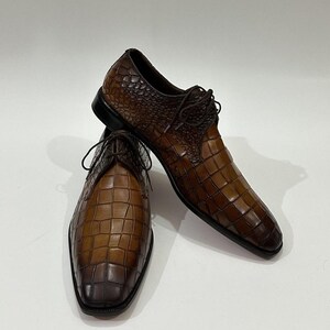 May include: A pair of brown leather dress shoes featuring a textured crocodile-embossed pattern with a gradient finish, dark brown laces, and a black sole. The shoes have a pointed toe design and a structured silhouette with visible stitching.