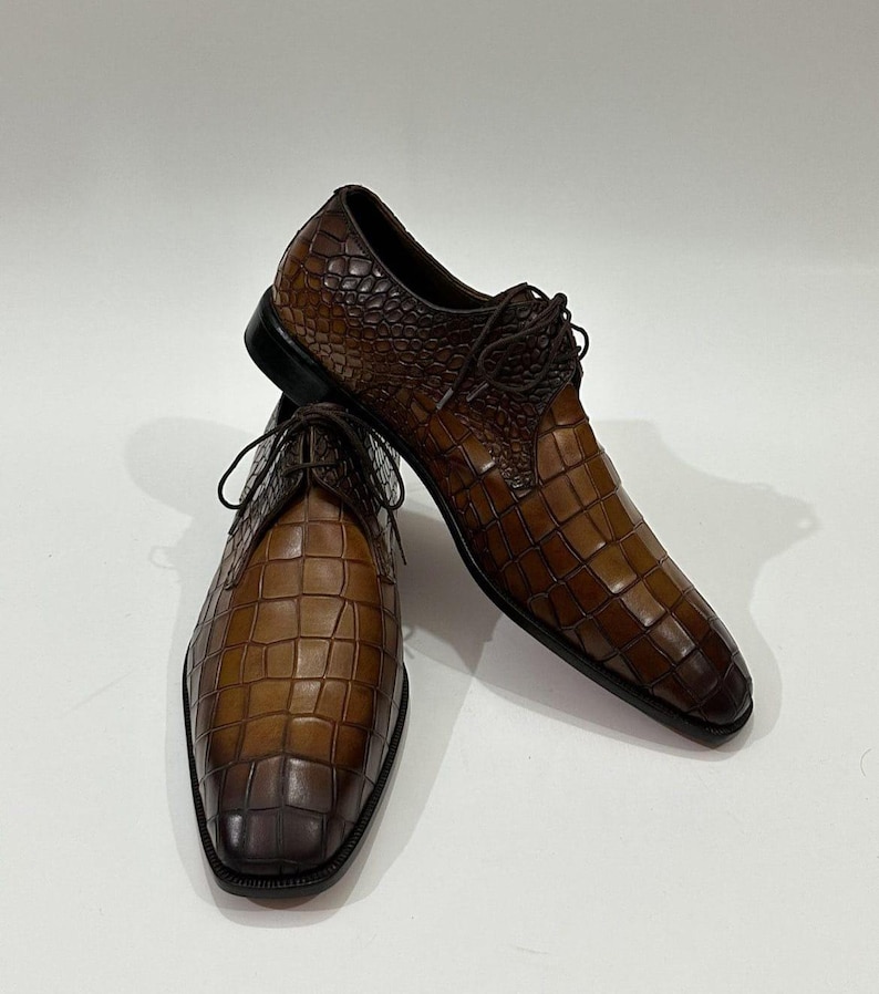 May include: A pair of brown leather dress shoes featuring a textured crocodile-embossed pattern with a gradient finish, dark brown laces, and a black sole. The shoes have a pointed toe design and a structured silhouette with visible stitching.