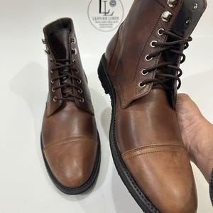 May include: A pair of brown leather lace-up boots with a cap toe design, dark brown laces, and silver-toned metal eyelets and speed hooks. A circular logo with the text LEATHER LORDS is visible in the background between the two boots.