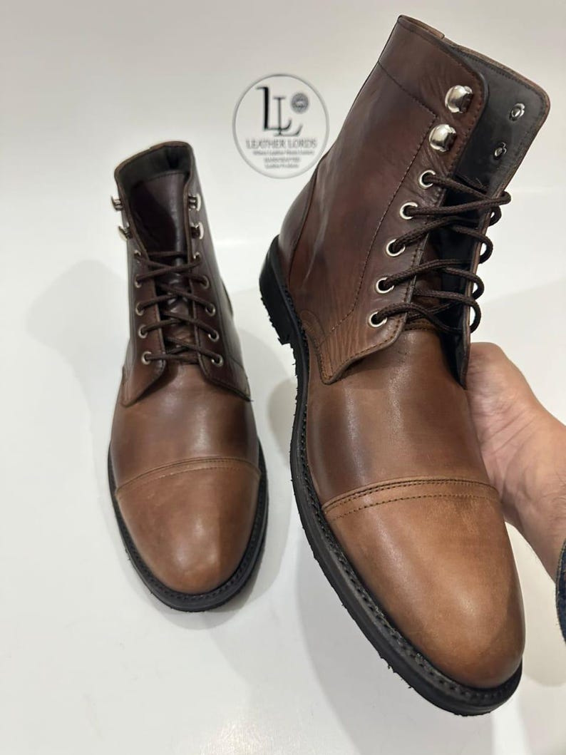 May include: A pair of brown leather lace-up boots with a cap toe design, dark brown laces, and silver-toned metal eyelets and speed hooks. A circular logo with the text LEATHER LORDS is visible in the background between the two boots.