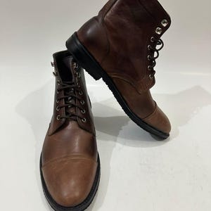 May include: A pair of brown leather lace-up boots featuring a cap toe design, dark brown laces, silver-toned metal eyelets, and a thick black rubber sole. The boots have a smooth finish and a structured ankle-high silhouette.