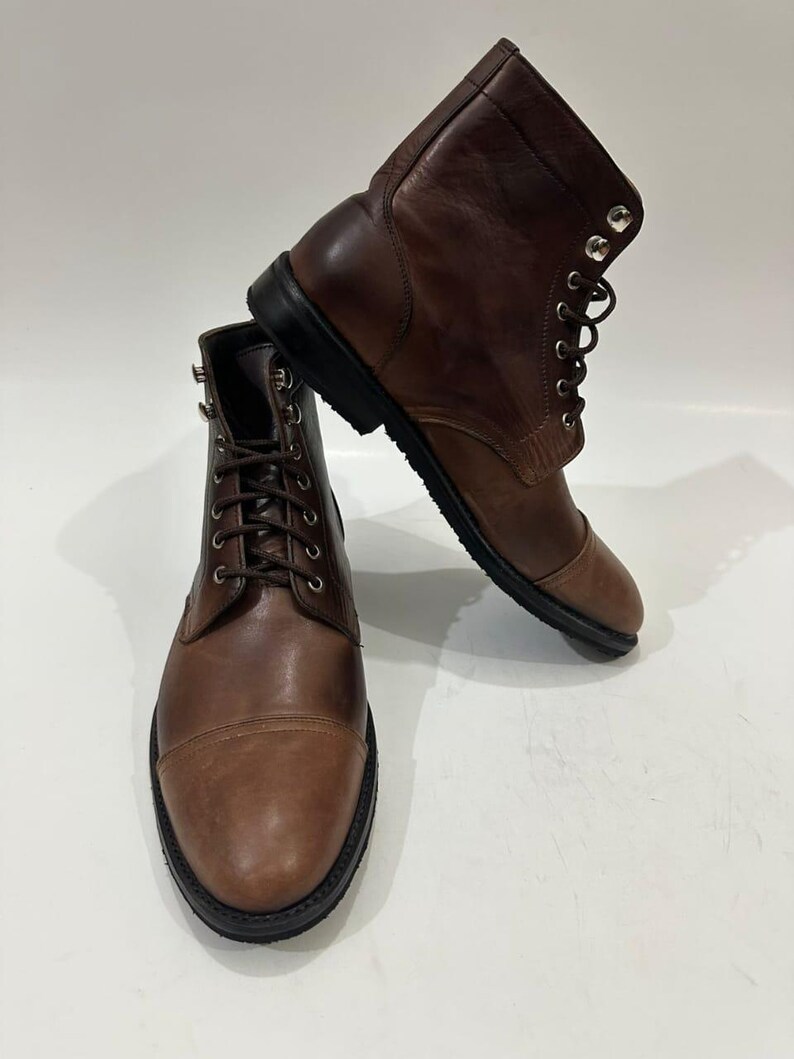 May include: A pair of brown leather lace-up boots featuring a cap toe design, dark brown laces, silver-toned metal eyelets, and a thick black rubber sole. The boots have a smooth finish and a structured ankle-high silhouette.