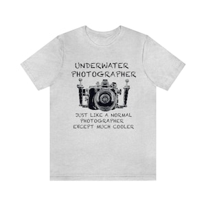 以下が含まれることがあります： 「UNDERWATER PHOTOGRAPHER」の文字と、防水ハウジングに入ったカメラの黒いイラストがプリントされたライトグレーのTシャツ。「JUST LIKE A NORMAL PHOTOGRAPHER EXCEPT MUCH COOLER」の文字が下にあります。
