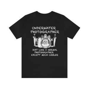 以下が含まれることがあります： 黒いTシャツ。白い文字と水中カメラのグラフィック。テキストはカメラ画像の上に「UNDERWATER PHOTOGRAPHER」、下に「JUST LIKE A NORMAL PHOTOGRAPHER EXCEPT MUCH COOLER」と書かれています。