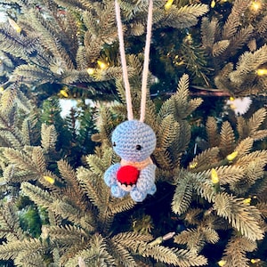 May include: A handmade blue crochet octopus Christmas ornament with a red heart, hanging from a cream-colored string. The octopus has a tan neck and is set against a backdrop of a green Christmas tree with small lights.