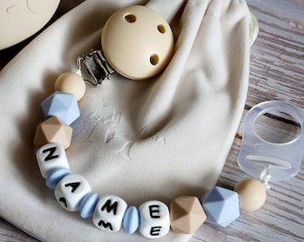 Può includere: Un mordedor per bambini in silicone blu e beige con il nome "NAME" scritto in lettere bianche. Il mordedor ha un clip per fissarlo ai vestiti e viene fornito con una piccola borsa di stoffa bianca.