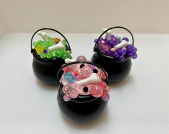 May include: Three small black cauldrons filled with colorful, sparkling concoctions. Each cauldron has a black handle and is filled with different colored beads, bone-shaped pieces, and other decorative elements, perfect for Halloween.