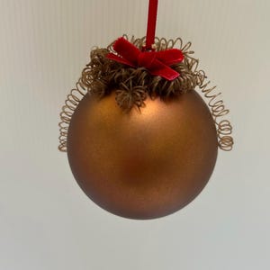 May include: A bronze-colored Christmas ornament with a red velvet bow and a decorative, curly, gold-colored trim. The ornament hangs from a red ribbon, perfect for holiday decorating.