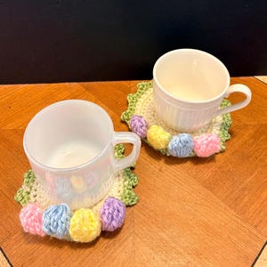 May include: Two white ceramic cups sit on handmade crochet coasters. The coasters are cream-colored with a green scalloped edge and adorned with colorful yarn pom-poms in pink, blue, yellow, and purple. The cups have a pearlescent sheen.
