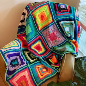 Könnte beinhalten: Eine bunte Häkeldecke mit einem quadratischen Muster in den Farben Grün, Blau, Rot, Gelb, Orange, Lila und Schwarz. Die Decke liegt über einem braunen Sessel.