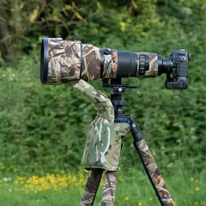 May include: A professional camera with a large telephoto lens, mounted on a tripod. The lens and tripod legs are covered in a camouflage pattern. The background is a blurred green field and trees.