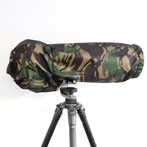 May include: A camouflage-patterned lens cover is attached to a tripod. The cover is made of a waterproof material and has a drawstring closure.