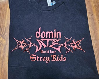 May include: Black t-shirt with a red graphic design that says "dominate" with a stylized font and "Stray Kids" underneath. The graphic also includes the words "World Tour" in a smaller font.
