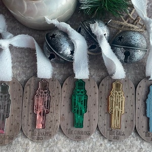 May include: Five wooden ornaments shaped like nutcrackers with different coloured mirrored finishes. The ornaments are decorated with stars and have the names of families and the year 2023 printed on them. The ornaments are hanging from white ribbon.