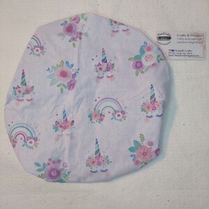 May include: A light purple fabric item featuring a pattern of unicorns, rainbows, and floral designs in pink, purple, and blue. The item has a tag that says "Crafts & Designs".