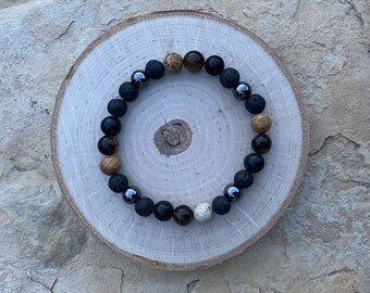 May include: A beaded bracelet with black, brown, and grey beads. The bracelet is on a wooden surface.