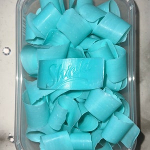 May include: A clear plastic container filled with light blue soap curls. The soap curls are in a variety of shapes and sizes. The container has a label that says "Shield" on it.