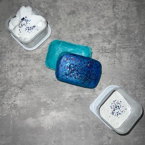 Puede incluir: Tres recipientes con tapas blancas, cada uno contiene un producto de jab&oacute;n azul y blanco diferente. El primer recipiente contiene un jab&oacute;n espumoso blanco con purpurina azul. El segundo recipiente contiene una barra de jab&oacute;n azul con purpurina azul y el texto "Shweel" en &eacute;l. El tercer recipiente contiene un polvo blanco con purpurina azul.