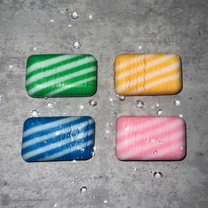 Puede incluir: Cuatro jabones rectangulares en verde, amarillo, azul y rosa, cada uno con rayas diagonales blancas. Los jabones están dispuestos sobre una superficie gris con gotas claras dispersas.