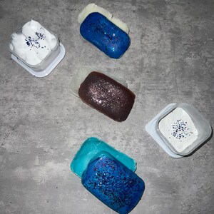 Puede incluir: Cuatro recipientes con tapas blancas y purpurina azul en el interior. Cada recipiente tiene una pastilla de jab&oacute;n a juego azul, morado o blanco con purpurina azul en la parte superior.