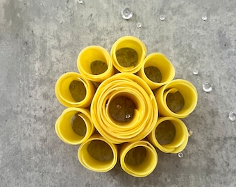 Op de afbeelding: Een gele papieren bloem gemaakt van opgerolde cirkels van papier. De bloem ligt op een grijze achtergrond.