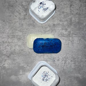 Puede incluir: Tres recipientes con polvo blanco y purpurina azul. El recipiente superior tiene una sustancia blanca y esponjosa, el recipiente del medio tiene una barra azul s&oacute;lida y el recipiente inferior tiene un polvo blanco fino.