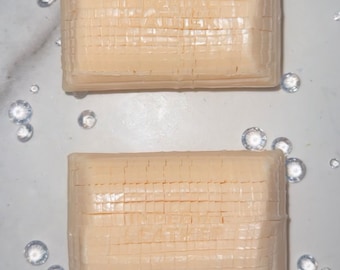 May include: Two rectangular bars of ivory-coloured soap with a grid pattern on the surface. The soap bars are on a white marble background.
