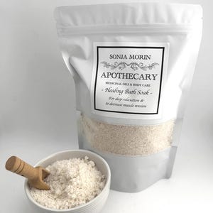 Peut inclure: Un sac refermable blanc rempli d'un bain moussant blanc. Le sac porte une étiquette sur laquelle est inscrit "SONJA MORIN APOTHECARY MEDICINAL OILS & BODY CARE - Healing Bath Soak - For deep relaxation & to decrease muscle tension". Un petit bol blanc avec une cuillère en bois est rempli de bain moussant.