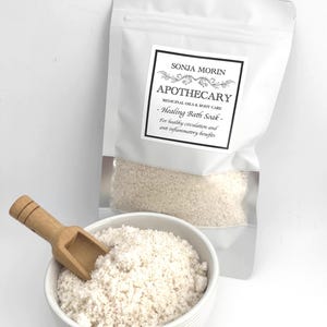 Puede incluir: Una bolsa blanca con una etiqueta que dice "SONJA MORIN APOTHECARY MEDICINAL OILS & BODY CARE - Healing Bath Soak - For healthy circulation and anti inflammatory benefits". La bolsa contiene un baño de inmersión blanco. Un tazón blanco con una cuchara de madera está lleno de baño de inmersión.