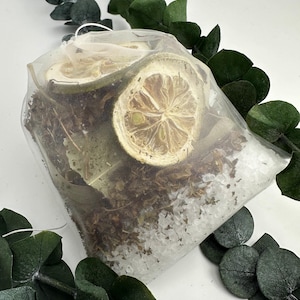 Puede incluir: Una bolsa de malla transparente llena de rodajas de limón secas, hierbas y sales de baño blancas. La bolsa está rodeada de hojas verdes de eucalipto.