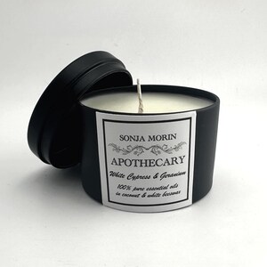 May include: A black candle jar with a white label that reads "SONJA MORIN APOTHECARY White Cypress & Geranium 100% pure essential oils in coconut & white beeswax".