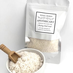 Puede incluir: Una bolsa blanca con una etiqueta que dice "SONJA MORIN APOTHECARY MEDICINAL OILS & BODY CARE - Healing Bath Soak - For deep relaxation & to decrease muscle tension". La bolsa contiene un baño de inmersión blanco y una pequeña pala de madera.