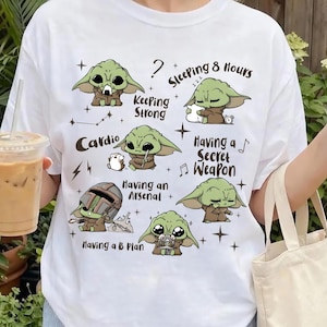 K&ouml;nnte beinhalten: Wei&szlig;es T-Shirt mit mehreren Illustrationen einer gr&uuml;nen Alien-Figur und S&auml;tzen wie &bdquo;Keeping Strong&ldquo;, &bdquo;Sleeping 8 Hours&ldquo; und &bdquo;Having a Secret Weapon&ldquo;. Das Wort &bdquo;Cardio&ldquo; ist ebenfalls auf dem T-Shirt aufgedruckt.