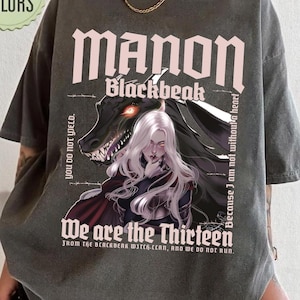 May include: Dark gray t-shirt featuring the text "MANON Blackbeak" above an illustration of a woman with white hair and a dragon. The shirt also includes the phrases "We are the Thirteen" and "You do not yield."