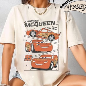 May include: Ivory-colored t-shirt featuring a graphic of Lightning McQueen from the movie Cars. The design includes three images of the character with the number 95, and the text "Lightning McQueen" and "Take the Lead."