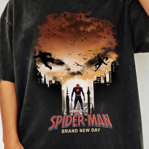 Może przedstawiać: Ciemnoszara koszulka z grafiką Spider-Mana. Projekt zawiera sylwetki postaci, krajobrazy miejskie oraz napis "SPIDER-MAN BRAND NEW DAY" w kolorze czerwonym i niebieskim. Widoczne jest r&oacute;wnież logo Marvel Studios.