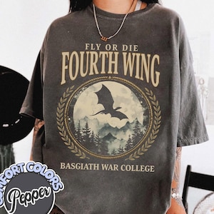 Puede incluir: Camiseta gris oscuro con el texto "FLY OR DIE FOURTH WING" sobre un gr&aacute;fico circular de una silueta de drag&oacute;n sobre un paisaje monta&ntilde;oso. El texto "BASGIATH WAR COLLEGE" est&aacute; debajo. La camiseta es de la marca Comfort Colors.