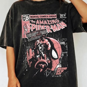 Peut inclure: T-shirt gris fonc&eacute; avec un design de couverture de bande dessin&eacute;e. Le design comprend le texte "The Amazing Spider-Man" en rose et blanc, des d&eacute;tails de bande dessin&eacute;e et une illustration de Spider-Man. Le t-shirt a un aspect vintage et us&eacute;.