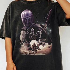 K&ouml;nnte beinhalten: Dunkelgraues T-Shirt mit einem Motiv aus einer Science-Fiction-Serie. Das Design zeigt eine Figur mit Helm, andere Charaktere und eine Sternenszene. Das T-Shirt hat einen Vintage-Look.