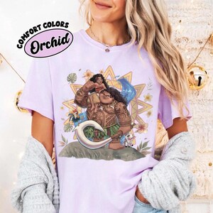 May include: A light purple t-shirt featuring a graphic print of the characters Moana and Maui from the film Moana, surrounded by tropical flowers, leaves, and a sun motif. The text COMFORT COLORS Orchid is printed in the upper left corner.