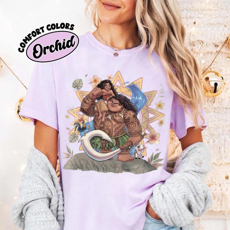 May include: A light purple t-shirt featuring a graphic print of the characters Moana and Maui from the film Moana, surrounded by tropical flowers, leaves, and a sun motif. The text COMFORT COLORS Orchid is printed in the upper left corner.
