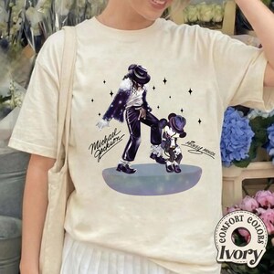 Pu&ograve; includere: Una t-shirt color avorio con una stampa grafica di Michael Jackson e Topolino in piedi su una piattaforma circolare. Entrambe le figure indossano fedora e abiti neri. Il testo Michael Jackson e Mickey Mouse &egrave; stampato in caratteri neri.