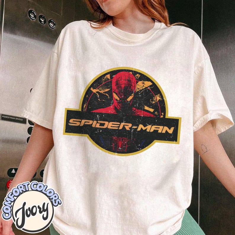 May include: An ivory-coloured short-sleeve t-shirt featuring a distressed graphic print on the chest. The design shows a red Spider-Man mask inside a yellow-bordered black circle, with the text SPIDER-MAN in yellow letters on a black banner.