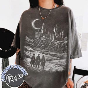 May include: A charcoal gray short-sleeve t-shirt featuring a white graphic print of a large castle on a hill under a crescent moon, with three figures in robes walking toward it. The shirt has a faded, washed-out appearance and a crew neckline.