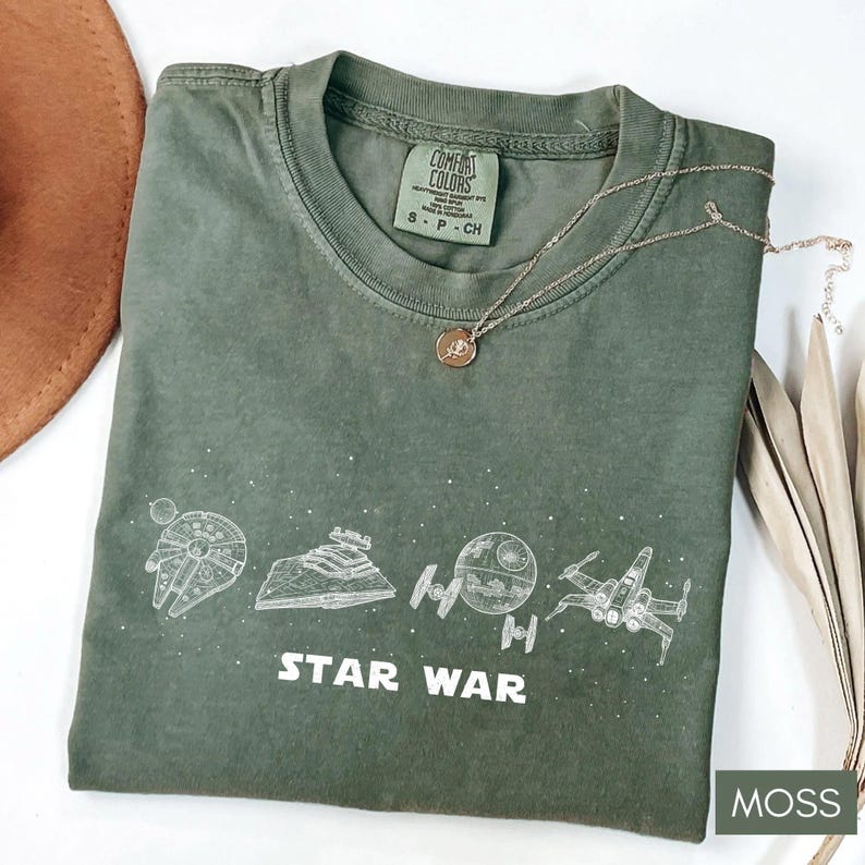 May include: A moss green t-shirt with a Star Wars graphic. The design includes illustrations of the Millennium Falcon, a Star Destroyer, the Death Star, and an X-wing fighter. The words "STAR WAR" are printed below the illustrations. A gold necklace rests on the shirt.