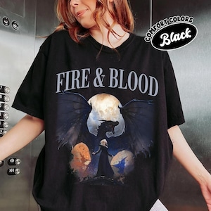 May include: A black short-sleeve t-shirt featuring the text FIRE & BLOOD in white capital letters. Below the text is a graphic illustration of a dark dragon with large wings spread wide in front of a full moon, with a figure standing in the foreground.