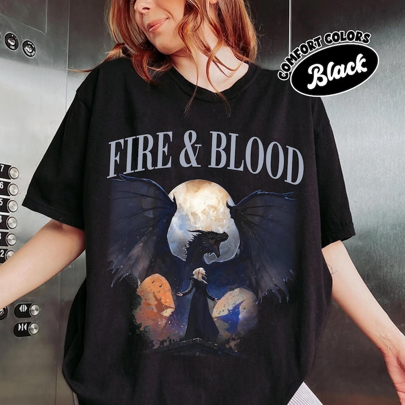 May include: A black short-sleeve t-shirt featuring the text FIRE & BLOOD in white capital letters. Below the text is a graphic illustration of a dark dragon with large wings spread wide in front of a full moon, with a figure standing in the foreground.