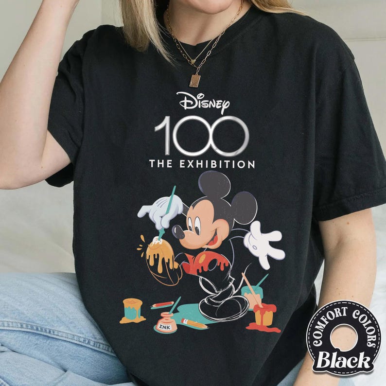 May include: Black t-shirt featuring Mickey Mouse painting, with the text "Disney 100 The Exhibition." The design includes paint pots and the "Comfort Colors" logo.